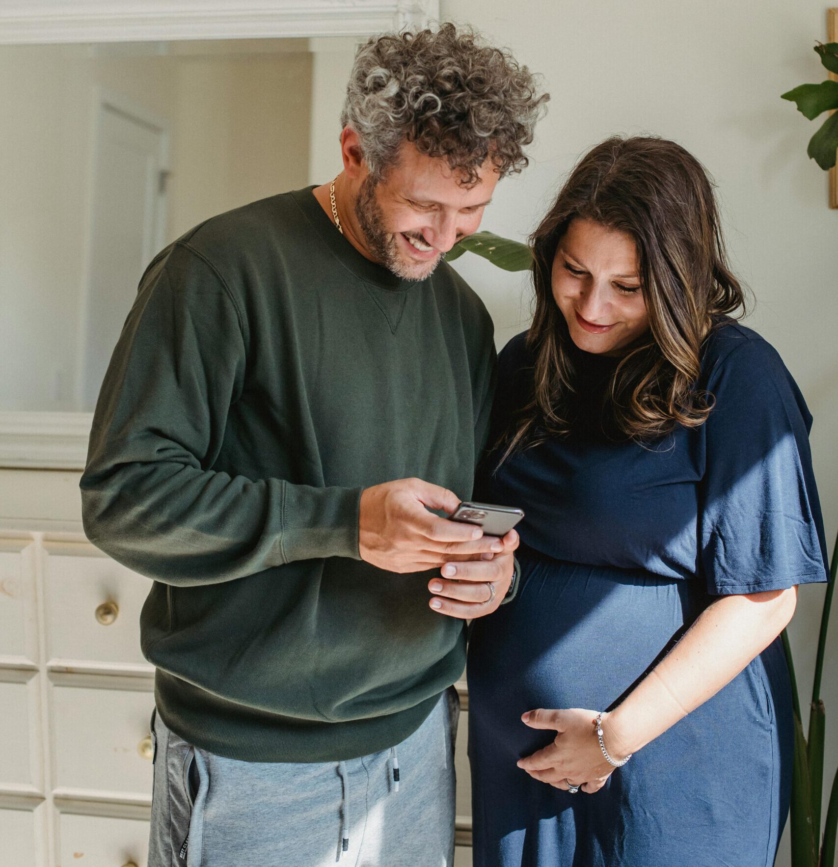 Cheerful adult male in casual clothes smiling and showing video on smartphone to smiling pregnant wife standing in light room on sunny day
